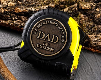 May include: Black and yellow tape measure with a bronze-colored face. The face reads "DAD" with the inscription "NO ONE MEASURES UP TO YOU LOVE BECKY & JASON" around the edge. The tape measure is on a gray surface with a piece of wood.