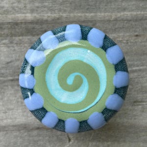 May include: A round, decorative knob with a hand-painted design. The center features a light blue spiral on a green background. The knob is encircled by a ring of light blue dots and a dark teal border.