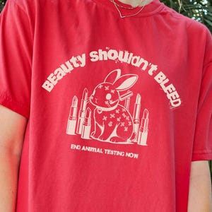 Puede incluir: Camiseta roja con un gráfico blanco de un conejo con lápiz labial y el texto "Beauty Shouldn't Bleed" y "End Animal Testing Now".
