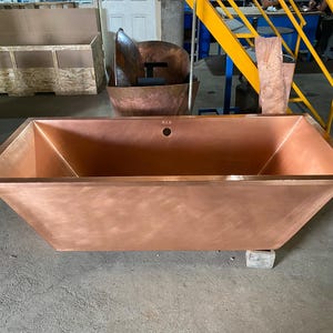 Puede incluir: Una bañera grande de color cobre con forma rectangular y lados angulados. La bañera tiene un orificio de drenaje y está sobre un suelo de hormigón. Otros objetos de cobre son visibles en el fondo.