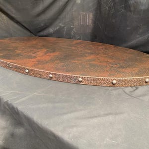 May include: Oval copper tabletop with a hammered texture and a dark patina. The edge is lined with small, silver-coloured rivets. The tabletop is approximately 122 cm long and 61 cm wide, suitable for a coffee table or similar furniture.