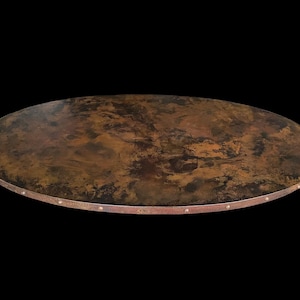 May include: Oval-shaped tabletop with a mottled brown and copper finish. The edge is trimmed with a thin, reddish-brown metal band and small, evenly spaced rivets. The table has a rustic, aged appearance.