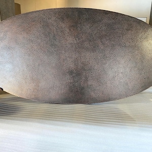 May include: Large, oval-shaped copper table top with a textured, hammered surface. The copper has a dark, mottled finish, with variations in color from light to dark brown. The table top is being held by two people.