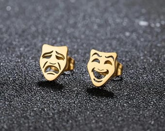 May include: A pair of gold-tone stainless steel earrings featuring a laughing and a crying face. The earrings are in the shape of a traditional theater mask.