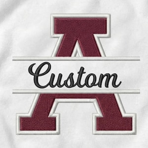 May include: A maroon embroidered letter A with a white outline, featuring a horizontal white bar across the center. The word Custom is stitched in black cursive script inside the white bar, all set against a white fabric background.