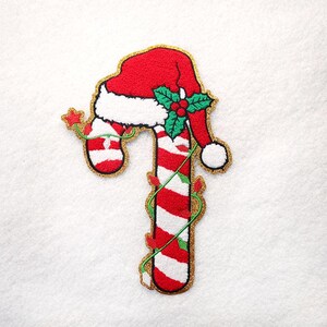 May include: An embroidered patch shaped like a red and white striped candy cane topped with a red Santa hat. The design features green holly leaves with red berries, a small red star, and a string of colorful Christmas lights wrapped around it.
