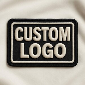 以下が含まれることがあります： 黒い背景に太字のサンセリフ体で「CUSTOM LOGO」という白い文字が刺繍された長方形のワッペン。ワッペンは太い白い枠と細い黒い外縁で囲まれており、すっきりとしたコントラストの高いデザインに仕上がっています。