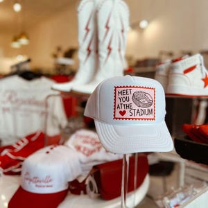 Könnte beinhalten: Eine weiße Trucker-Cap mit einer rechteckigen Grafik mit rotem Wellenrand. Die Grafik zeigt einen rosafarbenen Hintergrund, eine schwarze Strichzeichnung eines Stadions und den Text: MEET YOU AT THE STADIUM! mit einem roten Herzsymbol.