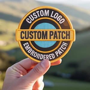 Peut inclure: Un écusson brodé circulaire avec un fond noir et un centre bleu clair. Le texte CUSTOM LOGO est arqué en haut, EMBROIDERED PATCH est arqué en bas, et une bande horizontale au milieu porte l'inscription CUSTOM PATCH en lettres noires.