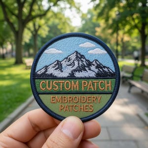 Puede incluir: Un parche bordado circular que muestra un paisaje de montaña con cielo azul y nubes blancas sobre un campo verde. El texto "CUSTOM PATCH" está bordado en marrón sobre una banda verde oscuro, con "EMBROIDERY PATCHES" debajo en marrón.