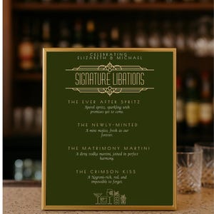 May include: A framed green sign with gold lettering and a gold border. The sign reads "Signature Libations" and lists four cocktail recipes: The Ever After Spritz, The Newly-Minted, The Matrimony Martini, and The Crimson Kiss.
