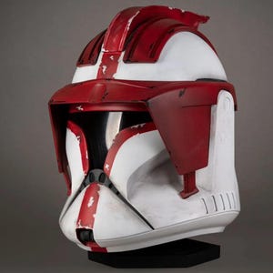 May include: A white and red helmet with a black visor. The helmet has a weathered appearance with red stripes and accents. The helmet is likely a prop or collectible item, possibly from a science fiction or fantasy theme.