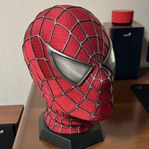 Op de afbeelding: Een Spider-Man masker replica, met een rood en zwart webpatroon en zilveren oogstukken. Het masker wordt getoond op een zwarte standaard, met het iconische superheldenontwerp. Een verzamelobject.