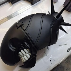 Puede incluir: Un casco negro con un diseño amenazante. Presenta puntas afiladas y una boca ancha que muestra dientes blancos. El diseño evoca un tema oscuro, futurista o de fantasía, con una estética imponente.