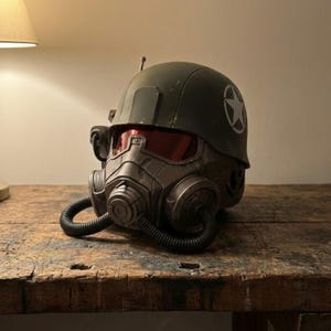 May include: A dark green military-style helmet with a white star emblem. The helmet features a respirator with a flexible black tube and red-tinted visor. The helmet is sitting on a wooden surface.