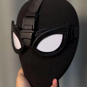 May include: Black mask with white eye lenses and a black strap. The mask features a rectangular device on top and is made of a flexible material. The eye lenses have a white mesh pattern.
