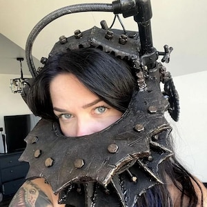 May include: A dark, metallic headpiece with a spiked collar and industrial-style components. The headpiece features bolts, gears, and a flexible tube, creating a steampunk or post-apocalyptic aesthetic. The design is predominantly black with metallic accents.