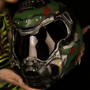 May include: A green and silver helmet with a dark visor. The helmet has red and yellow accents, with the text "R2-391" visible. The helmet is held in front of a dark background.