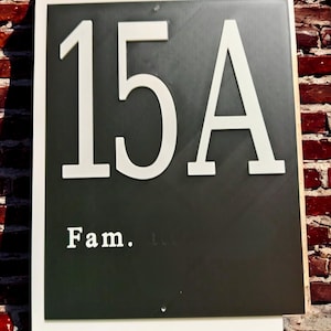 May include: A rectangular address sign with the number "15A" in white, bold font on a dark gray background. The word "Fam." is below the numbers. The sign is framed in white and mounted on a brick wall.