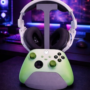Può includere: Cuffie da gioco bianche con padiglioni neri su un supporto grigio, sopra un controller Xbox verde e grigio. Il controller ha pulsanti verdi e una base grigia. Lo sfondo è una configurazione di computer sfocata.