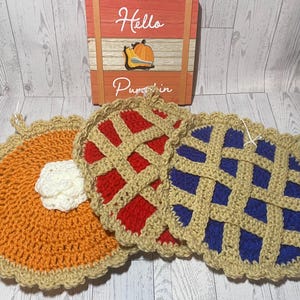 Könnte beinhalten: Gehäkelte Topflappen in Form von Kuchen. Einer ist orange mit einem weißen Klecks, ein anderer ist rot und der dritte ist blau. Die Topflappen haben ein beiges Gittermuster und einen gewellten Rand. Im Hintergrund befindet sich ein Holzschild mit der Aufschrift "Hello Pumpkin".