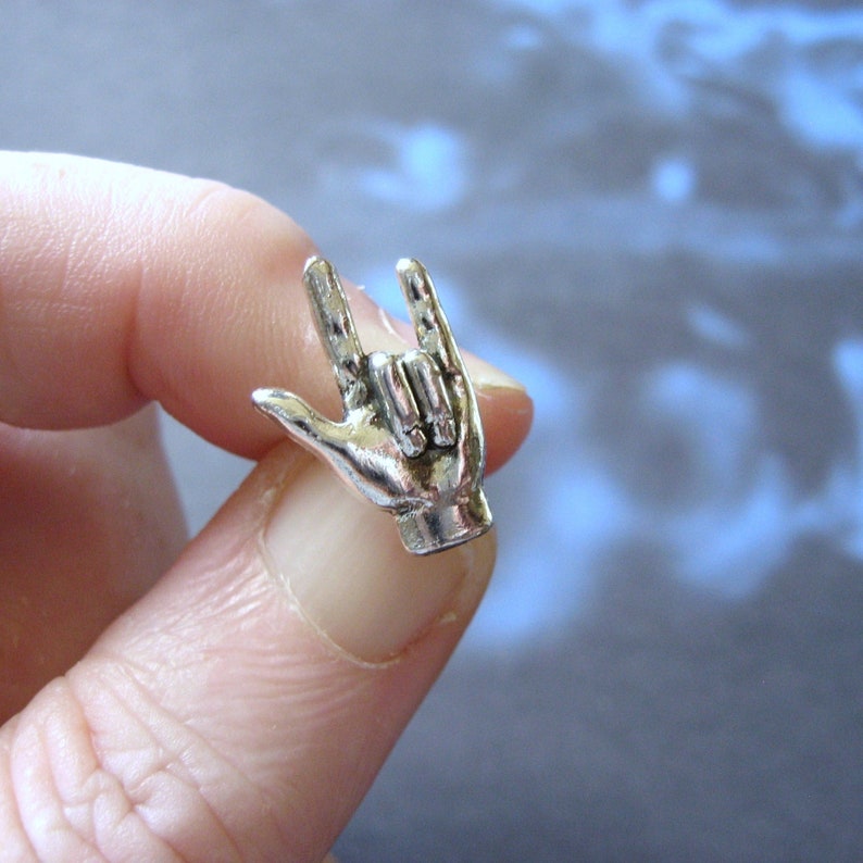 May include: A silver-colored metal charm in the shape of a hand making the 'I love you' sign. The charm is held between fingers, showcasing the detailed design of the hand gesture. The background is a blurred mix of blue and white.