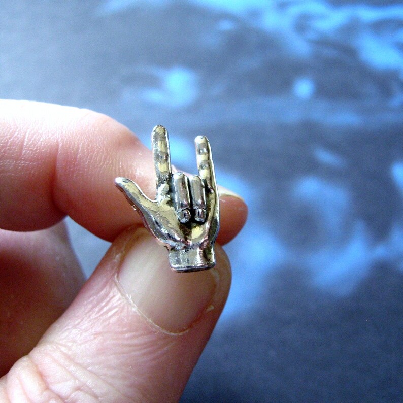 May include: A silver-colored metal charm in the shape of a hand making the 'rock on' gesture. The charm is held between fingers, showcasing the details of the hand's form. The background is a blurred blue and white.