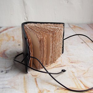 May include: A small, black leather-bound journal with a brown paper interior and a black cord closure. The journal is open and the pages are fanned out.
