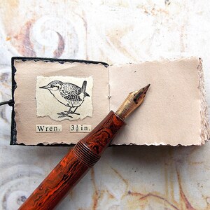 May include: A small black leather-bound journal with a brown and black pen on top of it. The journal is open to a page with a black and white illustration of a wren and the text "Wren. 3 1/2 in."