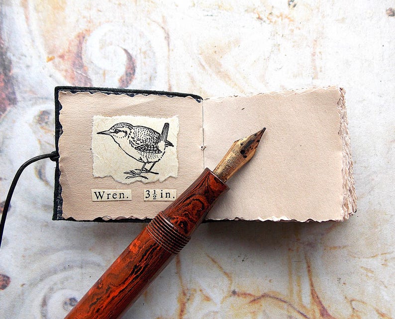 May include: A small black leather-bound journal with a brown and black pen on top of it. The journal is open to a page with a black and white illustration of a wren and the text "Wren. 3 1/2 in."