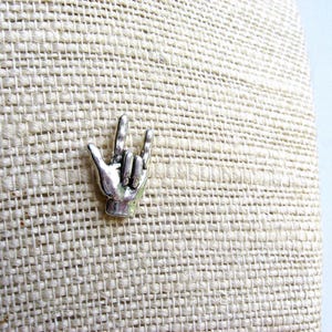 May include: A silver-colored metal pin in the shape of a hand making the 'I love you' gesture. The pin is set against a textured, beige fabric background. The pin is a small, detailed accessory.