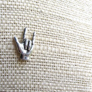 May include: A silver-colored metal pin in the shape of a hand making the 'I love you' sign. The pin is set against a textured, beige fabric background. The hand gesture is a popular symbol of affection and connection.