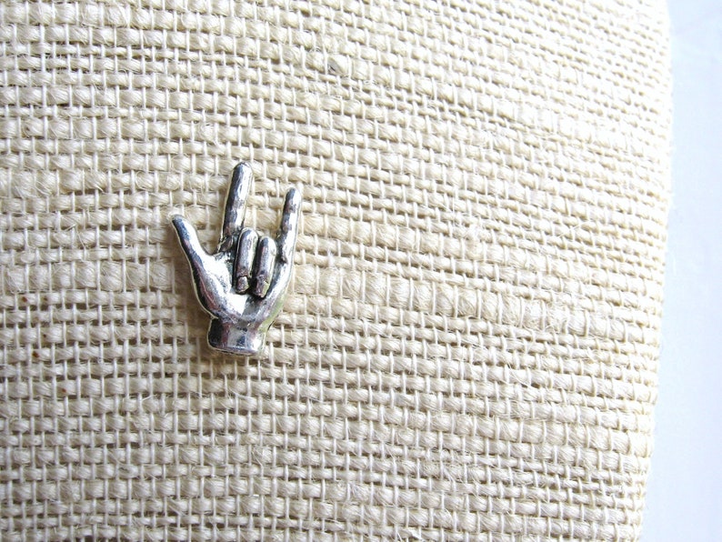 May include: A silver-colored metal pin in the shape of a hand making the 'I love you' sign. The pin is set against a textured, beige fabric background. The hand gesture is a popular symbol of affection and connection.