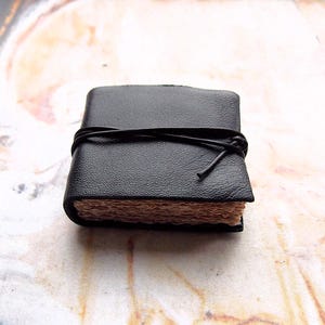 May include: A small black leather-bound journal with a brown paper interior and a black leather tie closure.