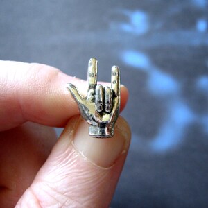 May include: A silver-colored metal charm in the shape of a hand making the 'rock on' gesture. The charm is detailed with lines and textures, and the fingers are slightly curved. The charm is held between fingers against a blurred background.