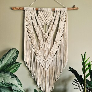 Może przedstawiać: Beigeowa wisząca ozdoba ścienna wykonana techniką makramy z geometrycznym wzorem i frędzlami. Ozdoba jest przymocowana do naturalnego drewnianego gałęzi.