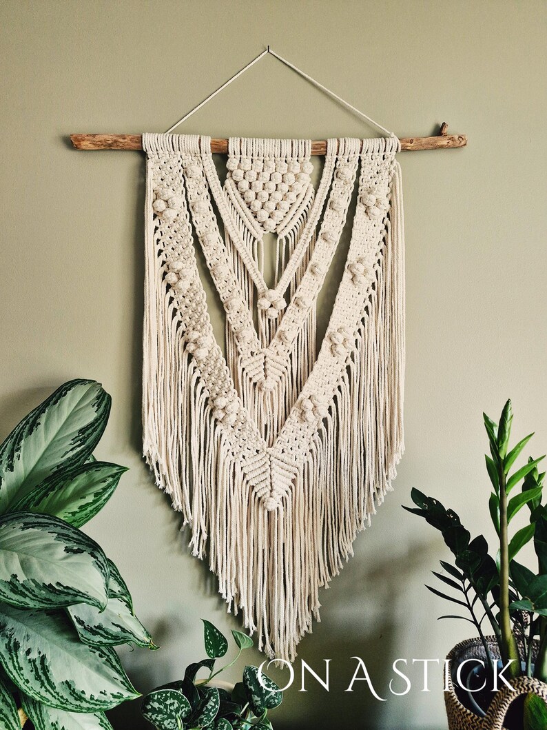 Może przedstawiać: Beigeowa wisząca ozdoba ścienna wykonana techniką makramy z geometrycznym wzorem i frędzlami. Ozdoba jest przymocowana do naturalnego drewnianego gałęzi.