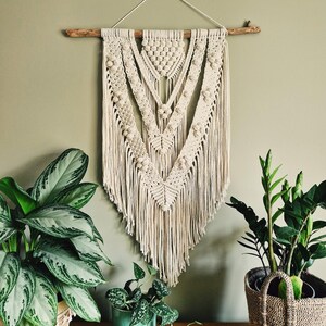 Może przedstawiać: Duży, beżowy wiszący gobelin z makramą o geometrycznym wzorze i frędzlami. Gobelin jest przymocowany do naturalnego drewnianego gałęzi i jest prezentowany na tle zielonej ściany.