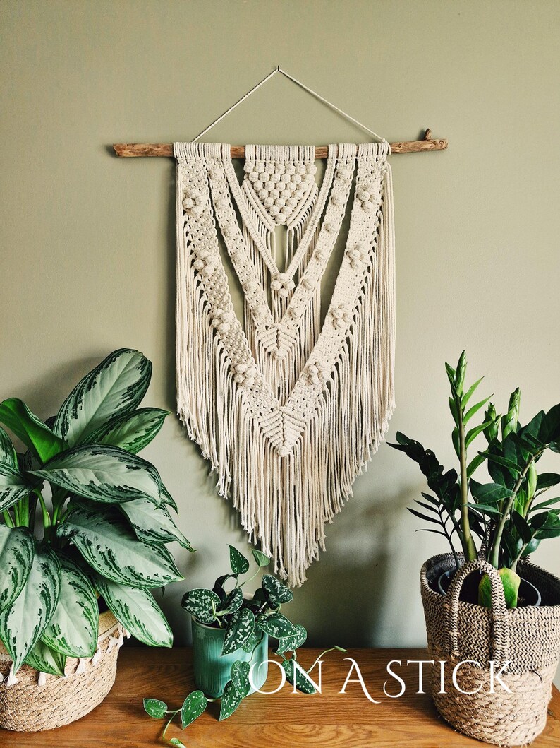 Może przedstawiać: Duży, beżowy wiszący gobelin z makramą o geometrycznym wzorze i frędzlami. Gobelin jest przymocowany do naturalnego drewnianego gałęzi i jest prezentowany na tle zielonej ściany.