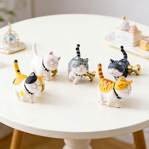 May include: Five decorative cat-shaped cabinet knobs in various colours, including white, orange, yellow, and grey, with black detailing. Each knob features a smiling cat face and a gold-coloured base. Ideal for adding a touch of whimsy.