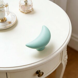 May include: A light blue crescent moon-shaped drawer pull on a white table. The drawer pull has a rounded base and a smooth, matte finish. The table has a decorative drawer pull and is part of a larger furniture set.