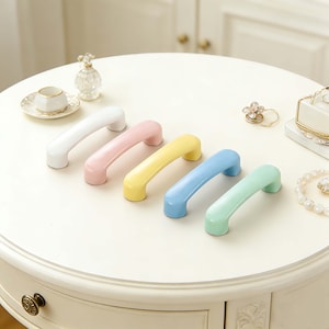 Puede incluir: Cinco tiradores de armario de colores blanco, rosa, amarillo, azul y verde están dispuestos sobre una mesa blanca. Cada tirador tiene un diseño curvo y arqueado. Los tiradores están hechos de un material liso y brillante.