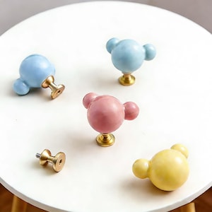 以下が含まれることがあります： 青、ピンク、黄色など、さまざまな色のキャビネットノブのコレクション。各ノブは、丸い本体と、クマの頭に似た2つの小さな丸い突起を備えています。ノブは金色のベースに取り付けられています。