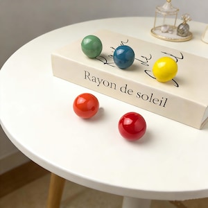 Könnte beinhalten: Eine Ansammlung farbenfroher, glänzender Kugeln in Grün, Blau, Gelb, Orange und Rot. Einige Kugeln liegen auf einem Buch mit dem Text "Rayon de soleil". Die Kugeln sind auf einem weißen Tisch angeordnet.