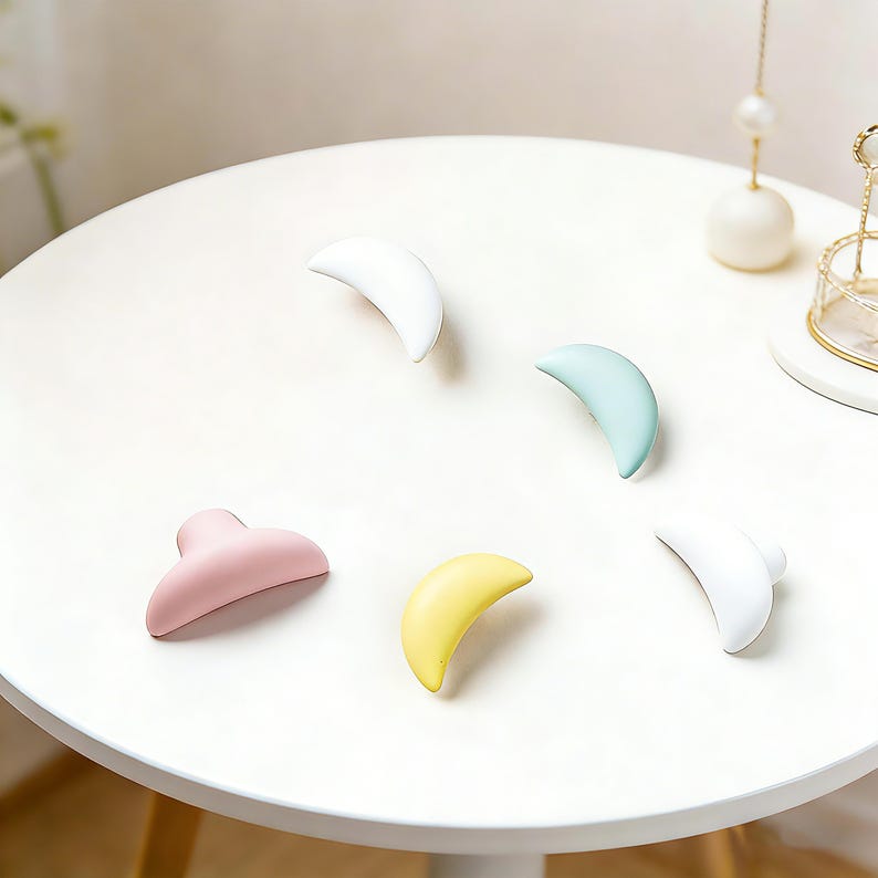 May include: Five pastel-colored hair claw clips arranged on a white surface. The clips are in shades of white, pink, yellow, and blue. The clips are crescent-shaped, with a curved design. The clips are made of a smooth, matte material.