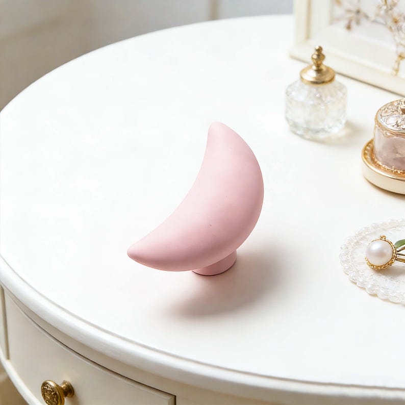 May include: A light pink crescent moon-shaped decorative object. The moon is on a small, round base. The object is on a white surface, with other decorative items in the background.