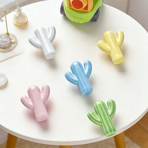 Op de afbeelding: Een verzameling kleine keramische cactusbeeldjes in verschillende pastelkleuren: wit, roze, blauw, geel en groen. De cactussen zijn gerangschikt op een wit oppervlak, met een kleine groene speelgoedauto op de achtergrond. Decoratieve items.