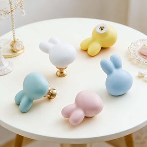 May include: Five pastel-colored bunny-shaped cabinet knobs. The knobs are in shades of white, yellow, blue, and pink. Each knob has a rounded body and two upright ears. The knobs are on a white table.