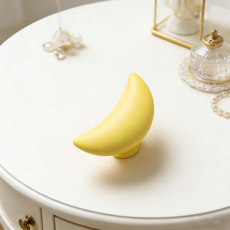 May include: A yellow crescent moon-shaped decorative object. The moon is smooth and sits on a small, round base. The object is on a white surface, with other decorative items in the background.