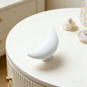 May include: A white crescent moon-shaped cabinet knob. The knob is mounted on a white surface, with other decorative items visible in the background. The knob is a decorative element for furniture.